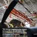 Military personnel are performing maintenance on a turboprop aircraft engine inside a large hangar facility. The image is overlaid with a motivational quote referencing mission support operations based in Georgia. The scene depicts active aircraft maintenance work with multiple technicians working simultaneously on the engine compartment.