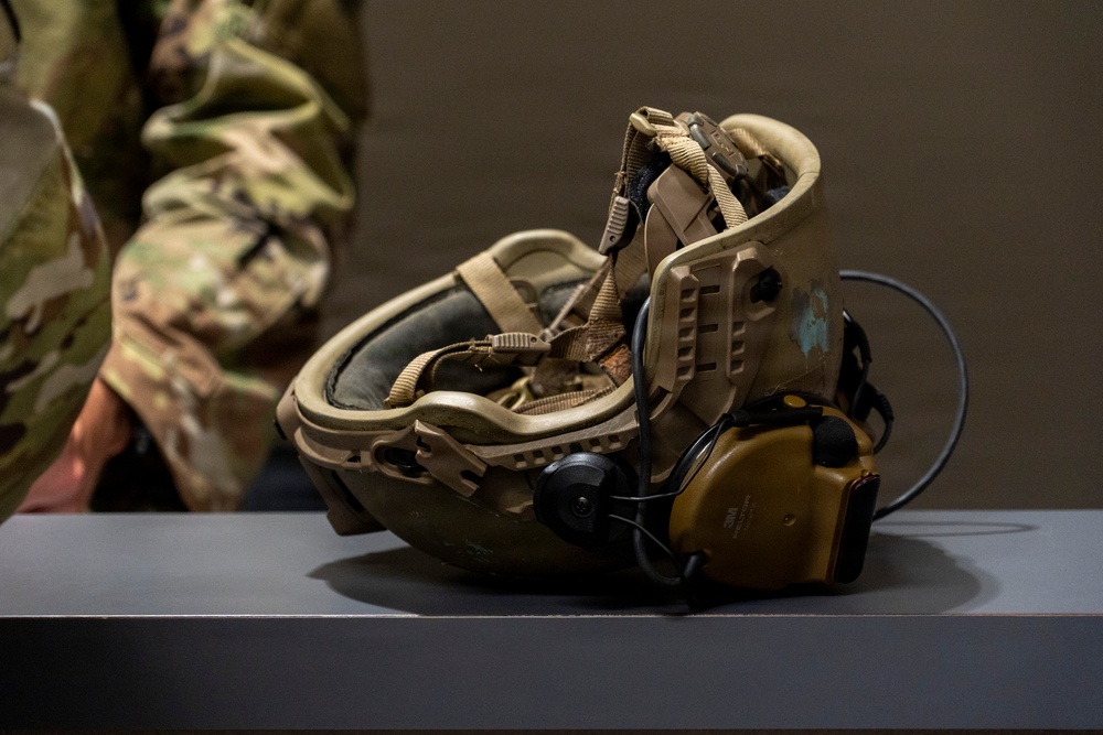 A military tactical helmet is placed upside down on a flat surface, revealing its interior padding, retention straps, and mounted communication headset. The helmet is equipped with electronic hearing protection/communication ear cups in a coyote tan/yellow color. A soldier in multicam/OCP camouflage uniform is partially visible in the background.