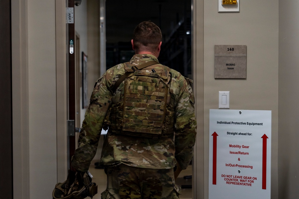 A military service member in OCP (Operational Camouflage Pattern) uniform is entering a doorway marked as Room 140, a MOBAG Issue facility. The individual is wearing a plate carrier/tactical vest and carrying additional gear. The scene depicts a military in/out-processing or equipment issue location.