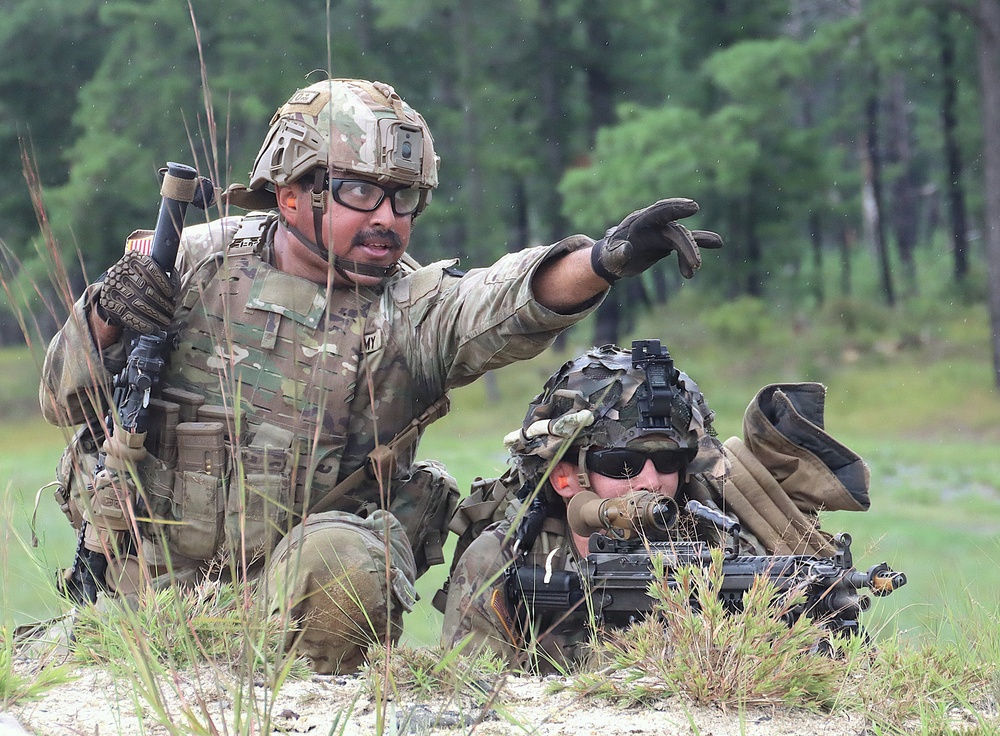 Two U.S. Army soldiers are engaged in a field training exercise in a wooded, grassy environment. One soldier is kneeling and pointing in a direction, apparently giving instructions or directing fire, while a second soldier is in a prone position behind a weapon system. Both are in full combat gear including OCP (Operational Camouflage Pattern) uniforms, tactical helmets, and body armor.