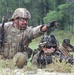 Two U.S. Army soldiers are engaged in a field training exercise in a wooded, grassy environment. One soldier is kneeling and pointing in a direction, apparently giving instructions or directing fire, while a second soldier is in a prone position behind a weapon system. Both are in full combat gear including OCP (Operational Camouflage Pattern) uniforms, tactical helmets, and body armor.