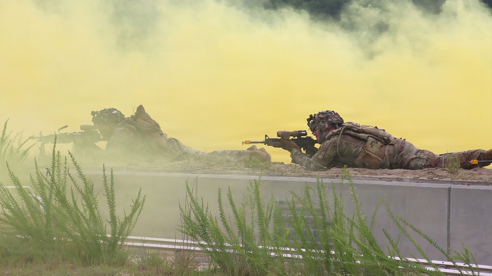 Two soldiers in full combat gear are in prone firing positions behind a concrete barrier, aiming rifles forward amid dense yellow-green smoke. The scene appears to be a military training exercise, indicated by the colored smoke grenades used for signaling or obscuration. The soldiers are wearing modern camouflage uniforms and tactical equipment consistent with contemporary Western military forces.
