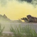 Two soldiers in full combat gear are in prone firing positions behind a concrete barrier, aiming rifles forward amid dense yellow-green smoke. The scene appears to be a military training exercise, indicated by the colored smoke grenades used for signaling or obscuration. The soldiers are wearing modern camouflage uniforms and tactical equipment consistent with contemporary Western military forces.