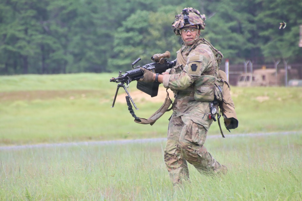 A soldier in full combat gear is moving across an open grassy field, carrying what appears to be an M249 Squad Automatic Weapon (SAW) in a ready position. The soldier is wearing Operational Camouflage Pattern (OCP) uniform, tactical helmet with accessories, protective eyewear, and a full load-bearing vest. The background suggests a military training range environment with tree lines and range infrastructure visible.