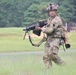 A soldier in full combat gear is moving across an open grassy field, carrying what appears to be an M249 Squad Automatic Weapon (SAW) in a ready position. The soldier is wearing Operational Camouflage Pattern (OCP) uniform, tactical helmet with accessories, protective eyewear, and a full load-bearing vest. The background suggests a military training range environment with tree lines and range infrastructure visible.