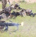 A line of soldiers in camouflage uniforms and helmet covers are in a prone firing position in a grassy field. At least one weapon (a light machine gun in the foreground) is visibly firing, indicated by a muzzle flash. The soldiers appear to be U.S. Army personnel, with at least one patch bearing an American flag visible.