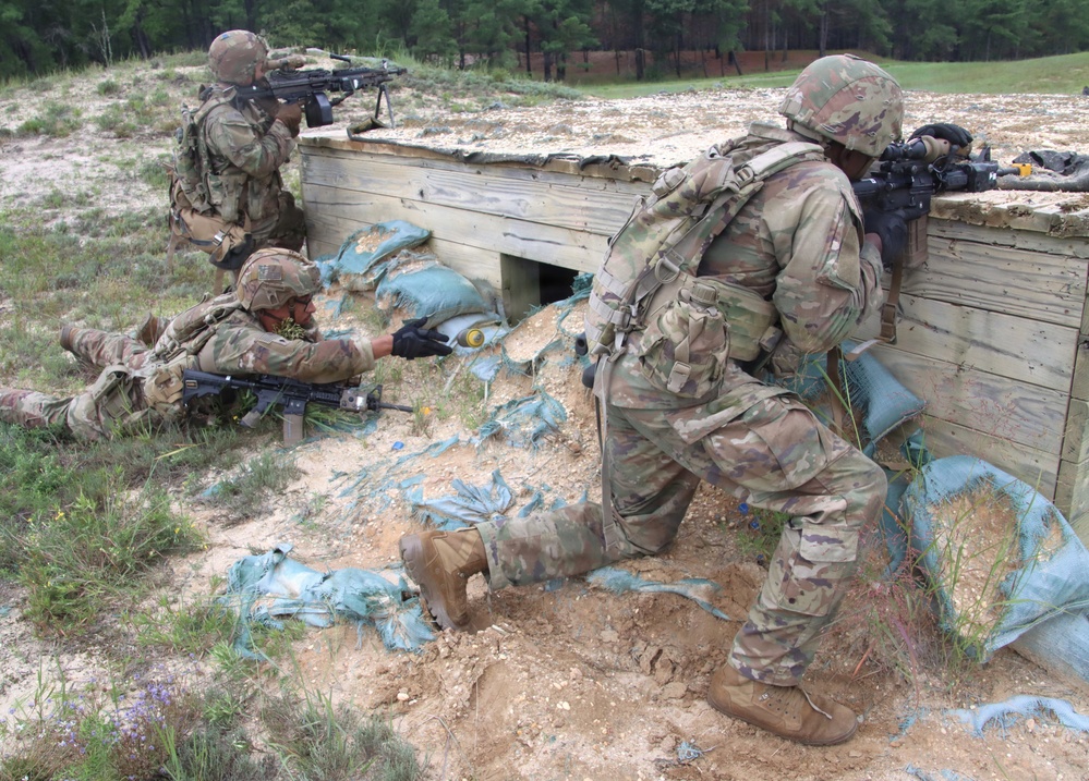 Three soldiers in MultiCam/OCP camouflage uniforms are conducting a tactical training exercise at what appears to be a military training range. They are positioned around a wooden fortified structure with sandbags, practicing defensive firing positions. One soldier is prone on the ground, one is kneeling, and one is standing, all with rifles raised or at the ready.