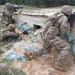 Three soldiers in MultiCam/OCP camouflage uniforms are conducting a tactical training exercise at what appears to be a military training range. They are positioned around a wooden fortified structure with sandbags, practicing defensive firing positions. One soldier is prone on the ground, one is kneeling, and one is standing, all with rifles raised or at the ready.