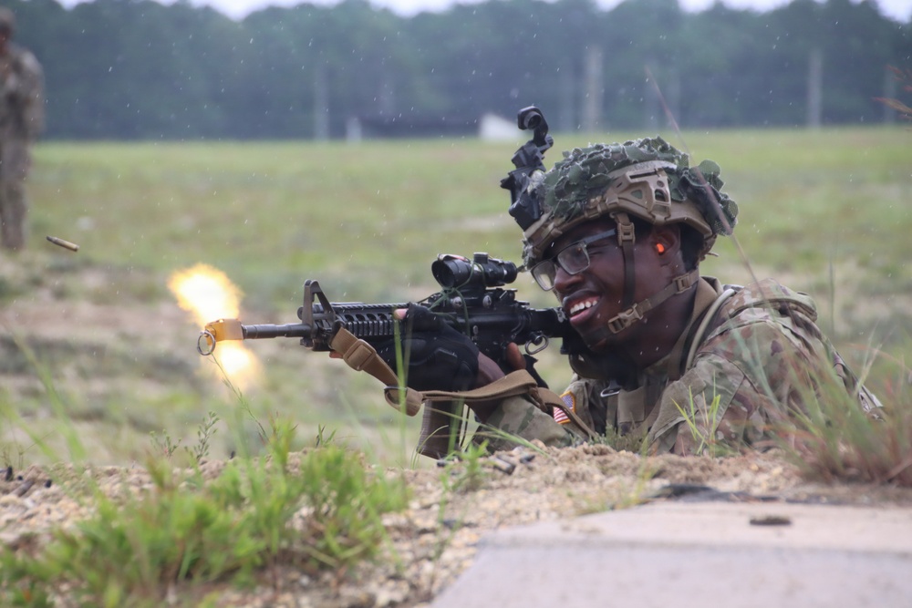 A U.S. Army soldier in full combat gear is firing an M4 carbine rifle from a prone position on a training range. A muzzle flash is clearly visible at the barrel, and an ejected brass casing can be seen mid-air to the left. The setting appears to be a military training exercise conducted in an open field environment.