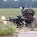 A U.S. Army soldier in full combat gear is firing an M4 carbine rifle from a prone position on a training range. A muzzle flash is clearly visible at the barrel, and an ejected brass casing can be seen mid-air to the left. The setting appears to be a military training exercise conducted in an open field environment.