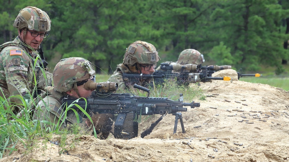 U.S. Army soldiers are conducting a live-fire training exercise at what appears to be a military range. Multiple soldiers in prone and crouching positions are firing weapons from a sandy berm, with spent casings visible on the ground. A soldier identified by name tape as 'YODER' is observing and supervising the exercise.