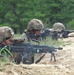 U.S. Army soldiers are conducting a live-fire training exercise at what appears to be a military range. Multiple soldiers in prone and crouching positions are firing weapons from a sandy berm, with spent casings visible on the ground. A soldier identified by name tape as 'YODER' is observing and supervising the exercise.