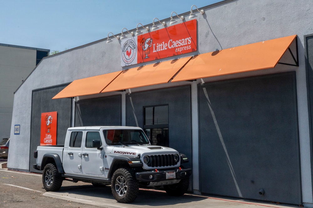 A white Jeep Gladiator Mojave pickup truck is parked in front of a Little Caesars Express restaurant location. The building features orange awnings and red Little Caesars branding signage, along with what appears to be a secondary business sign. The scene is photographed in clear daylight conditions.