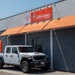 A white Jeep Gladiator Mojave pickup truck is parked in front of a Little Caesars Express restaurant location. The building features orange awnings and red Little Caesars branding signage, along with what appears to be a secondary business sign. The scene is photographed in clear daylight conditions.