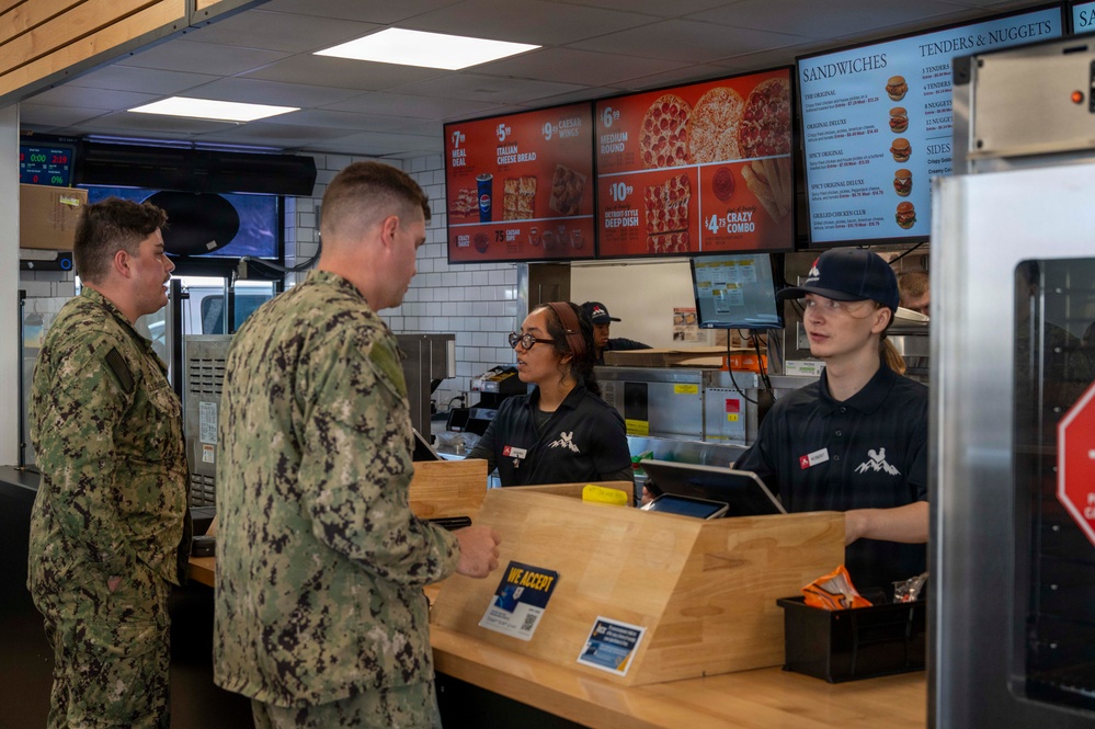 Two military personnel in camouflage uniforms are ordering food at a fast food counter, likely located on a military base. Two employees in black uniforms and caps are serving them at the counter. The restaurant appears to be a combination of Pizza Hut and another chain, based on the visible menu boards.