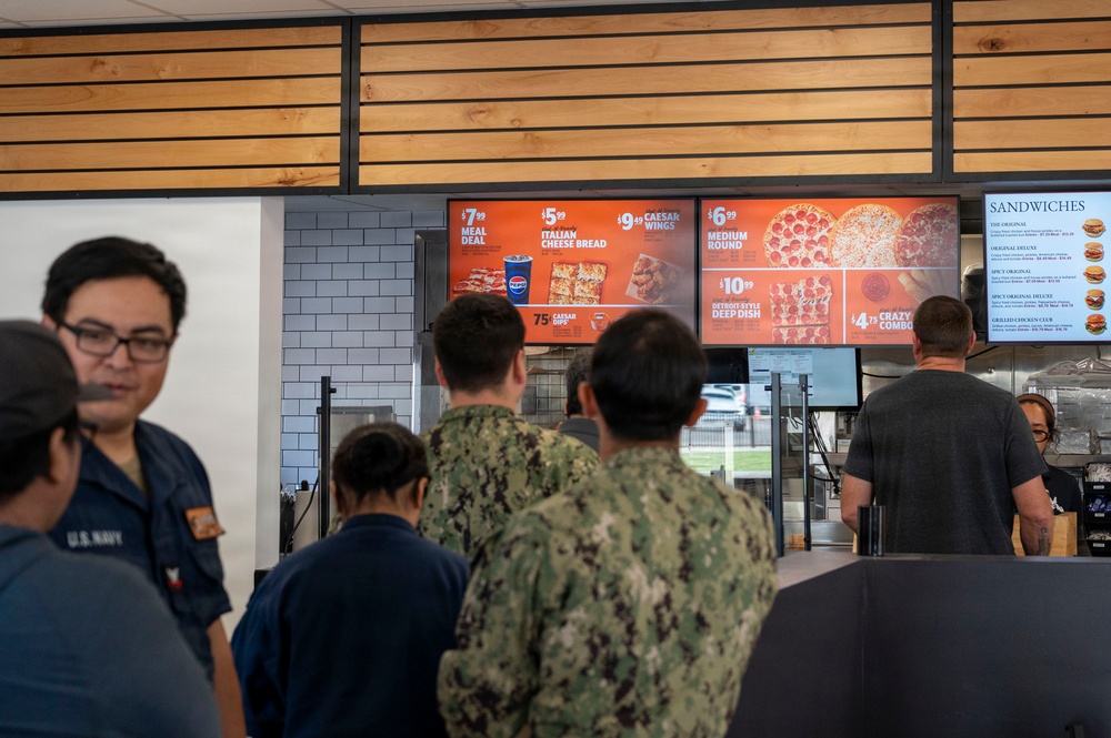 The image shows U.S. Navy personnel in uniform ordering food at what appears to be a Little Caesars pizza restaurant located on or near a military base. Several sailors in Navy Working Uniform (NWU) camouflage pattern are standing in line at the counter. Digital menu boards display pizza, wings, and sandwich options with visible pricing.