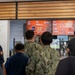 The image shows U.S. Navy personnel in uniform ordering food at what appears to be a Little Caesars pizza restaurant located on or near a military base. Several sailors in Navy Working Uniform (NWU) camouflage pattern are standing in line at the counter. Digital menu boards display pizza, wings, and sandwich options with visible pricing.