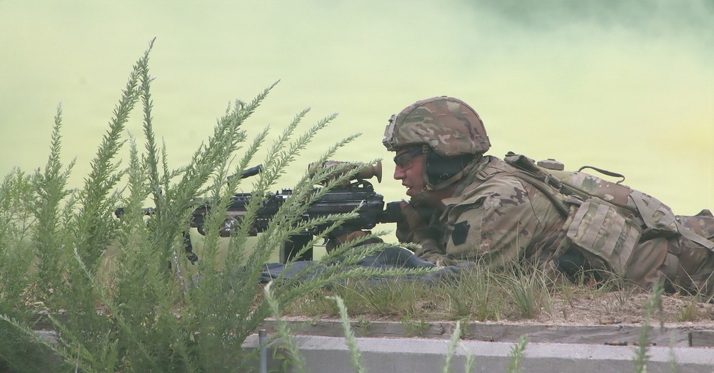 A soldier in full combat gear is in a prone firing position behind low vegetation and a concrete curb. The soldier is aiming a rifle forward while yellow-green smoke fills the background, suggesting a training exercise or tactical drill. The camouflage pattern and equipment appear consistent with modern U.S. Army MultiCam/OCP uniforms.