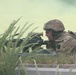 A soldier in full combat gear is in a prone firing position behind low vegetation and a concrete curb. The soldier is aiming a rifle forward while yellow-green smoke fills the background, suggesting a training exercise or tactical drill. The camouflage pattern and equipment appear consistent with modern U.S. Army MultiCam/OCP uniforms.