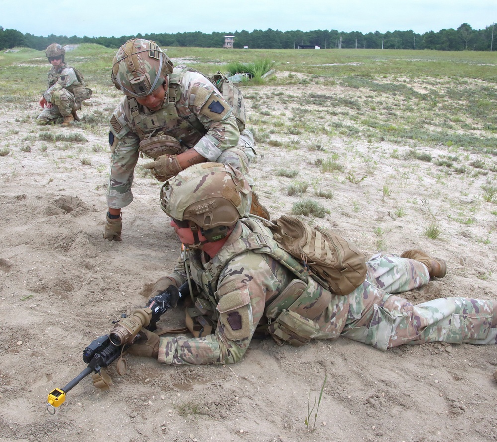 The image shows U.S. Army soldiers in Operational Camouflage Pattern (OCP) uniforms conducting a field training exercise on a sandy terrain range. One soldier is in the prone position holding a rifle while another soldier leans over them, appearing to provide instruction or correction on shooting technique. Additional soldiers are visible in the background in crouched positions.