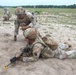 The image shows U.S. Army soldiers in Operational Camouflage Pattern (OCP) uniforms conducting a field training exercise on a sandy terrain range. One soldier is in the prone position holding a rifle while another soldier leans over them, appearing to provide instruction or correction on shooting technique. Additional soldiers are visible in the background in crouched positions.