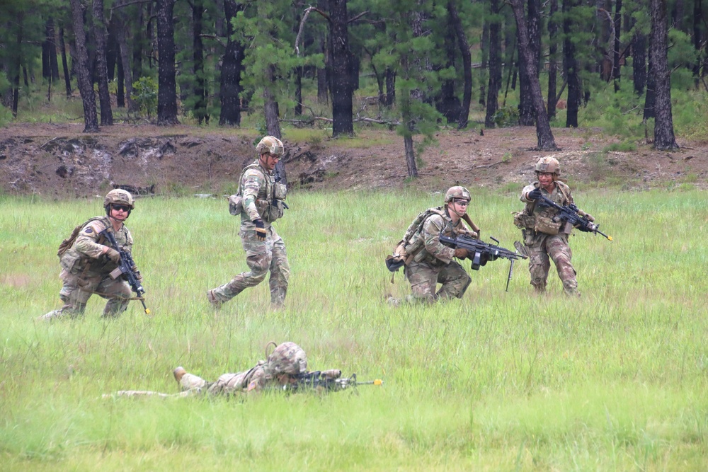 Four military personnel in camouflage uniforms and helmets are conducting a field training exercise in an open grassy area bordered by pine trees. The soldiers are in various tactical positions including prone, crouching, and standing while carrying rifles. The scene depicts a squad-level movement or assault drill consistent with U.S. Army training operations.