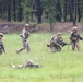 Four military personnel in camouflage uniforms and helmets are conducting a field training exercise in an open grassy area bordered by pine trees. The soldiers are in various tactical positions including prone, crouching, and standing while carrying rifles. The scene depicts a squad-level movement or assault drill consistent with U.S. Army training operations.