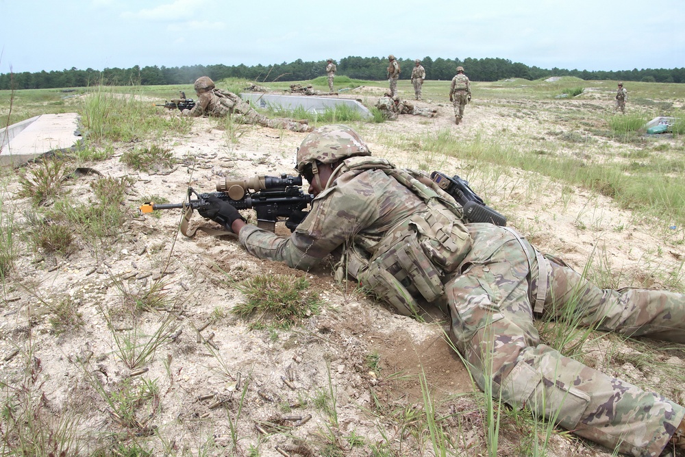 Military personnel in camouflage uniforms are conducting a field training exercise on a sandy, open terrain. The foreground soldier is in a prone firing position aiming a rifle equipped with an optic scope, while multiple other soldiers are visible in the background in various tactical positions. The setting appears to be a military training range with a treeline in the distance.