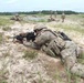 Military personnel in camouflage uniforms are conducting a field training exercise on a sandy, open terrain. The foreground soldier is in a prone firing position aiming a rifle equipped with an optic scope, while multiple other soldiers are visible in the background in various tactical positions. The setting appears to be a military training range with a treeline in the distance.