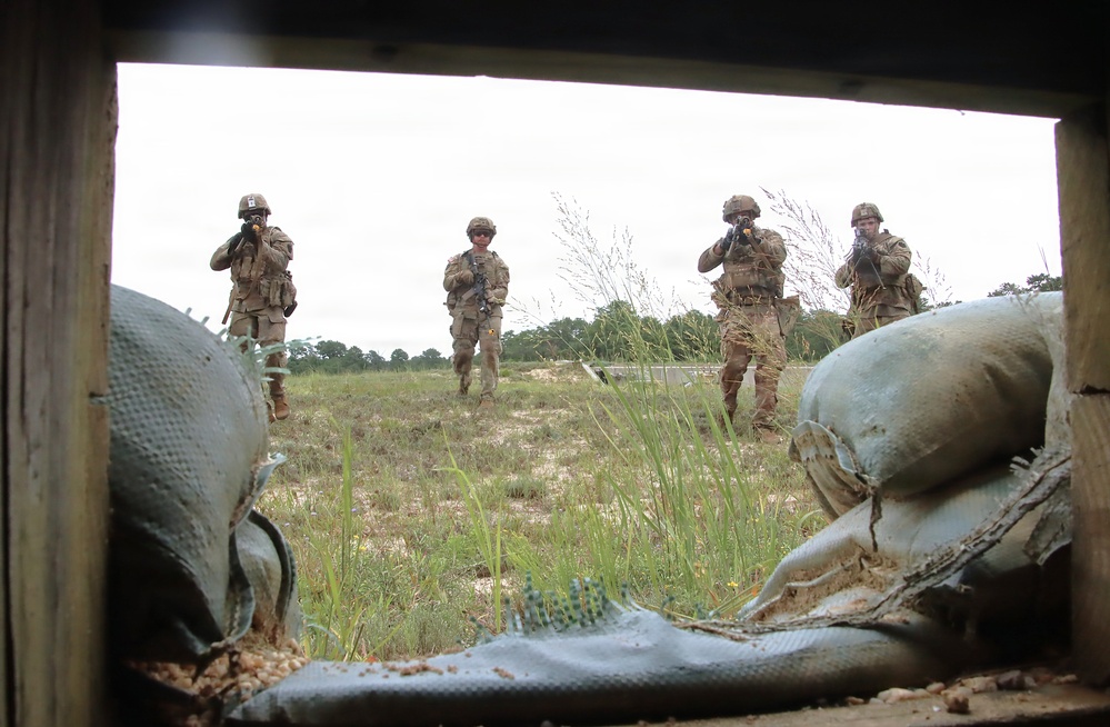 The image is taken from inside a fortified position through a window or opening framed by wooden planks, looking out at four soldiers in military camouflage uniforms advancing across an open grassy field. Three of the four soldiers are visibly raising their weapons in a tactical posture. The scene appears to be a military training exercise based on the controlled environment and visible sandbag fortifications.