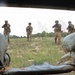 The image is taken from inside a fortified position through a window or opening framed by wooden planks, looking out at four soldiers in military camouflage uniforms advancing across an open grassy field. Three of the four soldiers are visibly raising their weapons in a tactical posture. The scene appears to be a military training exercise based on the controlled environment and visible sandbag fortifications.