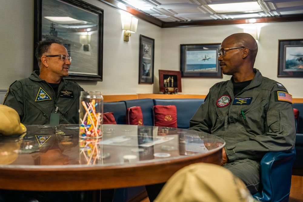 Two military personnel in olive/sage green flight suits are seated across from each other at a round glass-topped table, engaged in conversation. The setting appears to be an officer's lounge or ready room aboard a naval vessel, decorated with framed photographs of ships and aircraft. Both individuals wear unit patches and name tags on their flight suits.