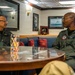 Two military personnel in olive/sage green flight suits are seated across from each other at a round glass-topped table, engaged in conversation. The setting appears to be an officer's lounge or ready room aboard a naval vessel, decorated with framed photographs of ships and aircraft. Both individuals wear unit patches and name tags on their flight suits.