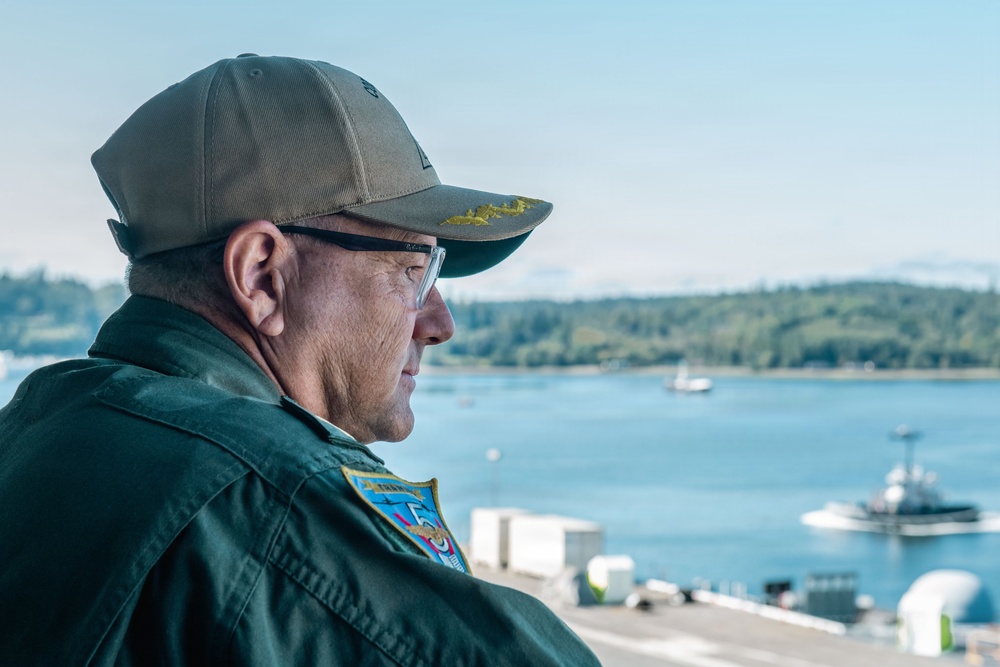 A middle-aged man wearing a dark green military-style flight suit and an olive/khaki baseball cap with gold embroidery on the brim is photographed in profile. He wears clear-framed glasses and has a colorful patch on his left shoulder. He appears to be overlooking a waterway with vessels and a tree-lined shoreline in the background.