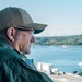 A middle-aged man wearing a dark green military-style flight suit and an olive/khaki baseball cap with gold embroidery on the brim is photographed in profile. He wears clear-framed glasses and has a colorful patch on his left shoulder. He appears to be overlooking a waterway with vessels and a tree-lined shoreline in the background.