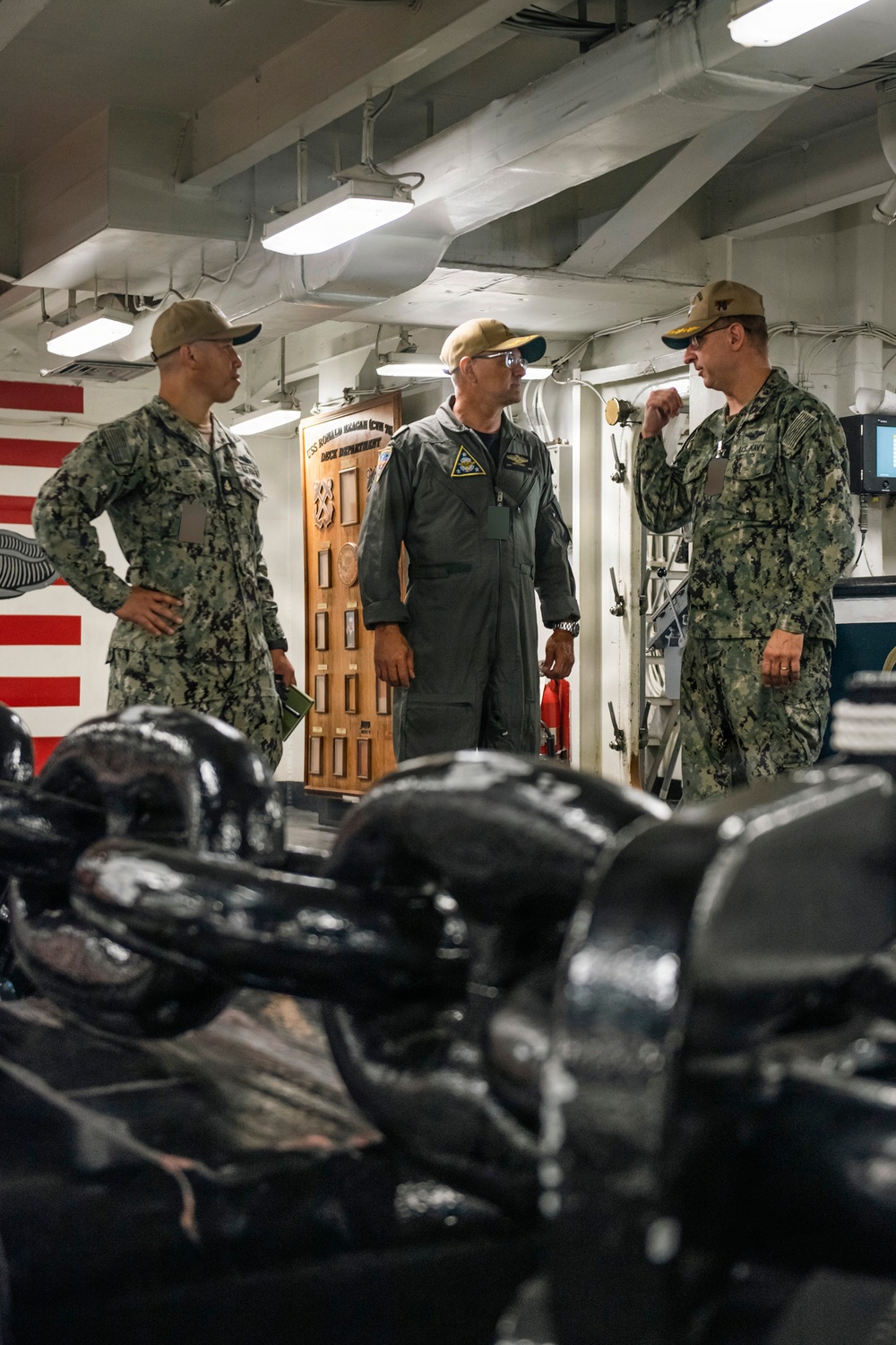 Three U.S. Navy personnel are engaged in conversation inside what appears to be a shipboard compartment, likely aboard the USS Ronald Reagan (CVN 76) based on the visible wooden placard in the background. Two individuals are wearing Navy Working Uniform (NWU) camouflage, while the center figure is wearing an olive-green flight suit with a squadron patch. Large black mooring lines or similar deck equipment are visible in the foreground.