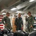 Three U.S. Navy personnel are engaged in conversation inside what appears to be a shipboard compartment, likely aboard the USS Ronald Reagan (CVN 76) based on the visible wooden placard in the background. Two individuals are wearing Navy Working Uniform (NWU) camouflage, while the center figure is wearing an olive-green flight suit with a squadron patch. Large black mooring lines or similar deck equipment are visible in the foreground.