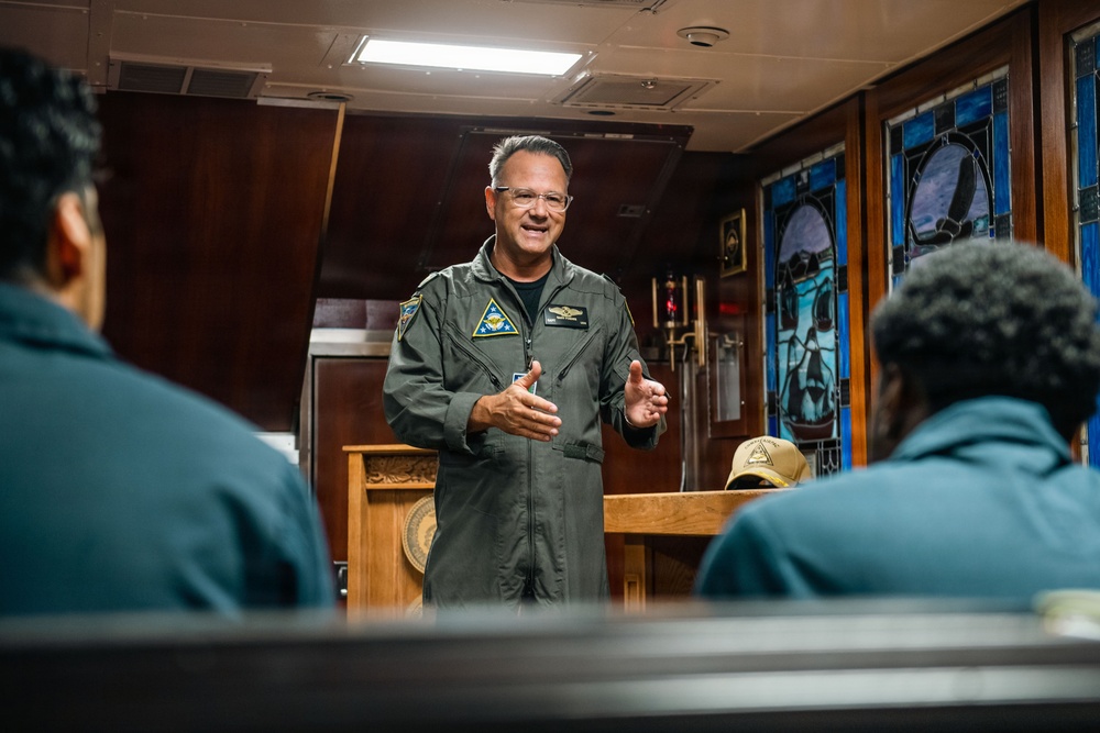 A military officer in an olive green flight suit is addressing a small group of seated individuals in what appears to be a chapel or ceremonial room aboard a naval vessel. The speaker is gesturing with both hands and smiling, suggesting an engaging presentation or briefing. Stained glass windows and wood-paneled walls are visible in the background.