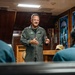 A military officer in an olive green flight suit is addressing a small group of seated individuals in what appears to be a chapel or ceremonial room aboard a naval vessel. The speaker is gesturing with both hands and smiling, suggesting an engaging presentation or briefing. Stained glass windows and wood-paneled walls are visible in the background.