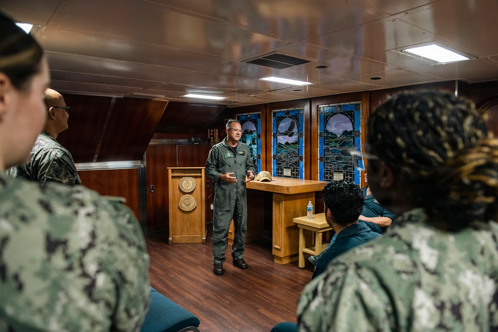 A military officer in a green flight suit is addressing a group of Navy personnel in what appears to be a ship's chapel or briefing room. The audience members are wearing Navy Working Uniform (NWU) camouflage pattern. The setting features distinctive stained glass windows, wood paneling, and a podium with a military seal.
