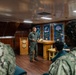 A military officer in a green flight suit is addressing a group of Navy personnel in what appears to be a ship's chapel or briefing room. The audience members are wearing Navy Working Uniform (NWU) camouflage pattern. The setting features distinctive stained glass windows, wood paneling, and a podium with a military seal.