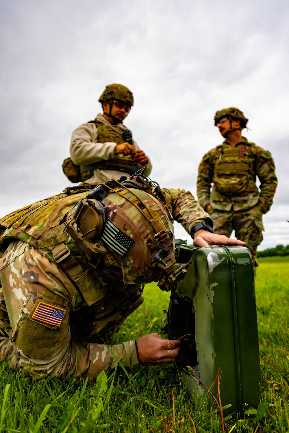 Three military personnel in MultiCam/OCP camouflage uniforms are working in an open grassy field under overcast skies. The foreground soldier is kneeling and manipulating a green military-style hard case or equipment box, while two other soldiers stand nearby observing. All personnel display US flag patches, indicating US Army affiliation.