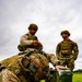 Three military personnel in MultiCam/OCP camouflage uniforms are working in an open grassy field under overcast skies. The foreground soldier is kneeling and manipulating a green military-style hard case or equipment box, while two other soldiers stand nearby observing. All personnel display US flag patches, indicating US Army affiliation.