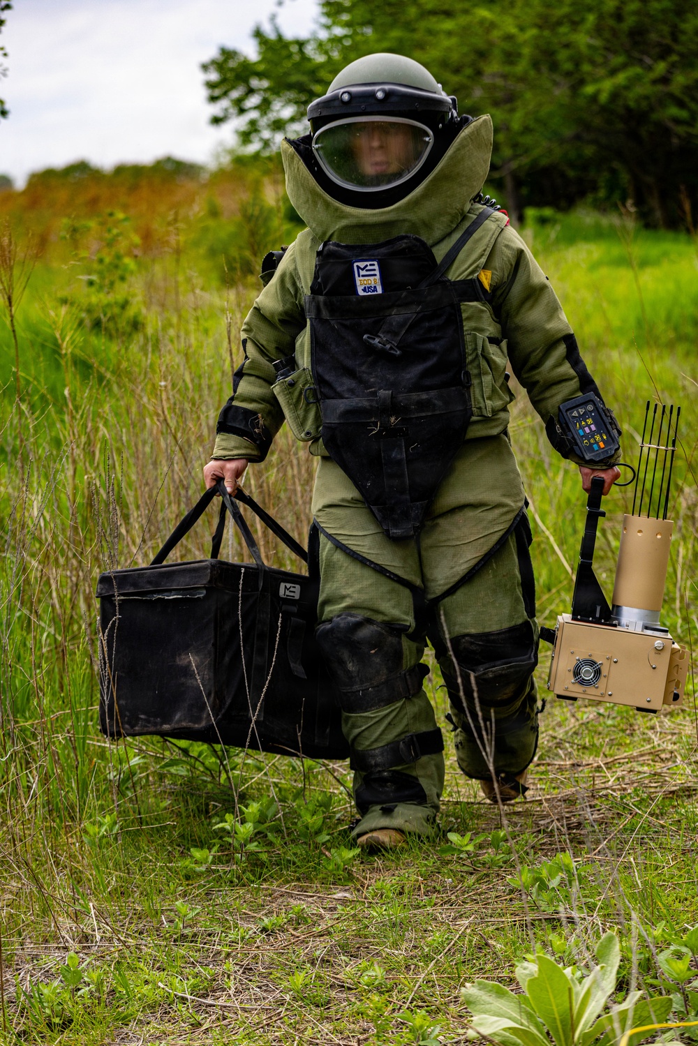 A person wearing a full EOD (Explosive Ordnance Disposal) bomb suit is walking through a grassy outdoor area. The individual is carrying an EOD tool bag in the left hand and what appears to be an electronic jamming or counter-IED device with multiple antennas in the right hand. The suit is marked with 'ME EOD II USA' branding, indicating a Med-Eng brand protective suit.