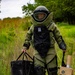 A person wearing a full EOD (Explosive Ordnance Disposal) bomb suit is walking through a grassy outdoor area. The individual is carrying an EOD tool bag in the left hand and what appears to be an electronic jamming or counter-IED device with multiple antennas in the right hand. The suit is marked with 'ME EOD II USA' branding, indicating a Med-Eng brand protective suit.
