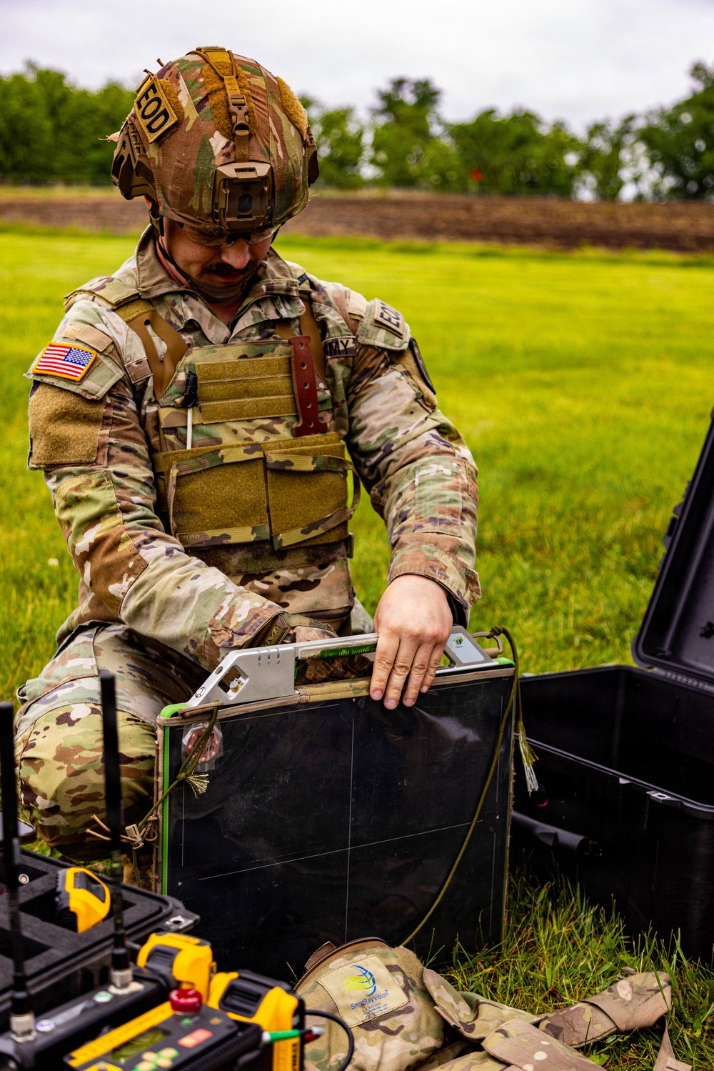 A U.S. Army EOD (Explosive Ordnance Disposal) technician in full OCP camouflage uniform and tactical gear is working with specialized equipment in an outdoor field setting. The soldier is handling what appears to be a ground-penetrating radar or detection panel device, with additional yellow-marked equipment cases visible in the foreground. The setting appears to be a training exercise or field operation on a grassy area.