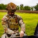 A U.S. Army EOD (Explosive Ordnance Disposal) technician in full OCP camouflage uniform and tactical gear is working with specialized equipment in an outdoor field setting. The soldier is handling what appears to be a ground-penetrating radar or detection panel device, with additional yellow-marked equipment cases visible in the foreground. The setting appears to be a training exercise or field operation on a grassy area.