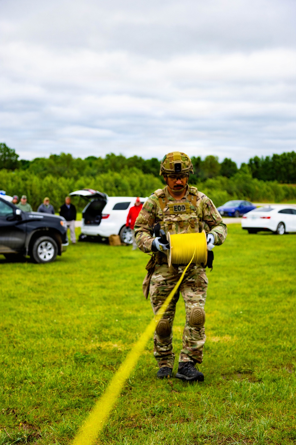 An EOD (Explosive Ordnance Disposal) technician in full tactical gear walks across a grassy field while unspooling a yellow wire or cord from a large reel. The individual is wearing multicam camouflage, a tactical helmet, body armor with an 'EOD' patch, gloves, and knee pads. Several observers and vehicles are visible in the background, suggesting this is a training exercise or demonstration.