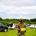 An EOD (Explosive Ordnance Disposal) technician in full tactical gear walks across a grassy field while unspooling a yellow wire or cord from a large reel. The individual is wearing multicam camouflage, a tactical helmet, body armor with an 'EOD' patch, gloves, and knee pads. Several observers and vehicles are visible in the background, suggesting this is a training exercise or demonstration.