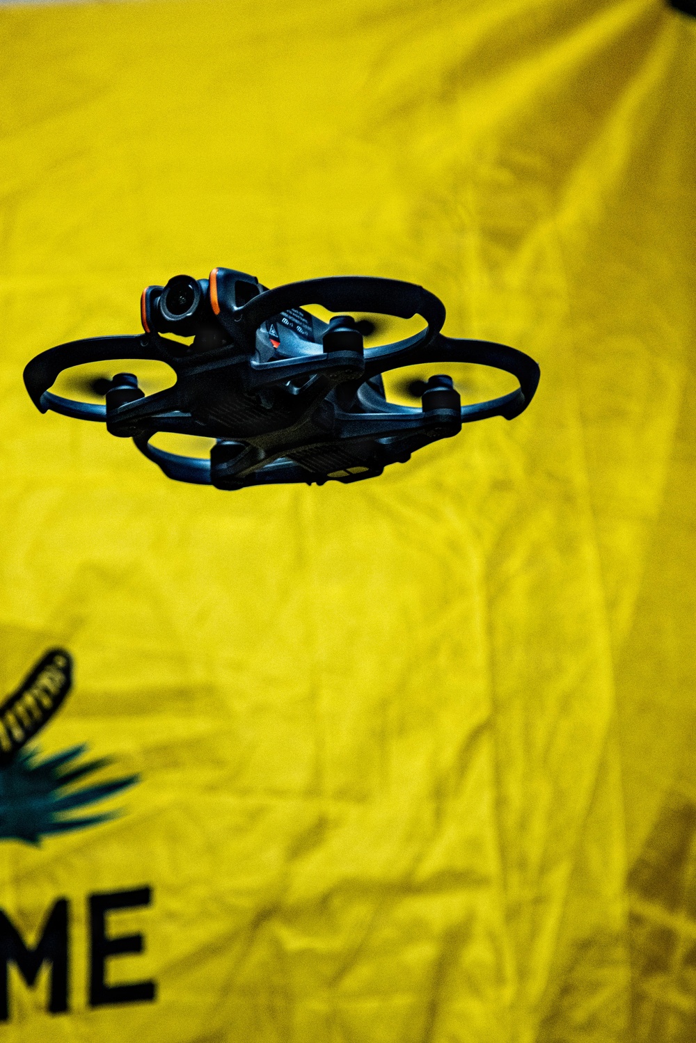 A small black FPV (First Person View) drone with ducted propeller guards is captured mid-flight against a bright yellow fabric backdrop. The drone features a forward-facing camera with an orange accent ring and enclosed rotor housings typical of indoor racing or cinematic micro drones. A partially visible graphic or logo appears in the lower-left corner of the yellow background.